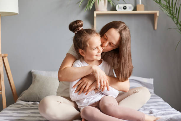 charmante petite fille félicitant maman pour la fête des mères, fille assise au lit avec maman, moment doux le matin, femme embrassant son enfant avec amour. - single mother one parent child kissing photos et images de collection