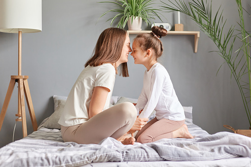 Side view portrait of brown haired woman and little girl, mother and daughter sitting in bed with closed eyes touching noses smiling playing together celebrating mother's day.