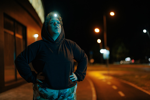 A young Caucasian overweight woman is standing with her hands on her hips, wearing a hoodie and a head lamp, resting after a night exercise session in the city.