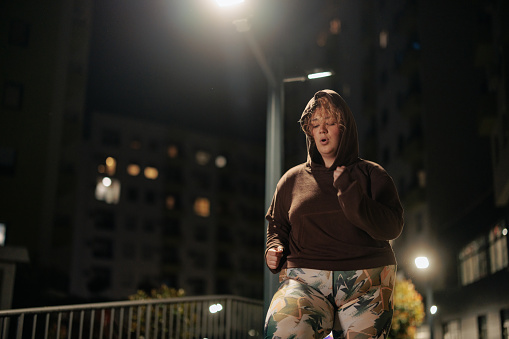 A young chubby Caucasian woman is outside in the city's urban area running in the evening.