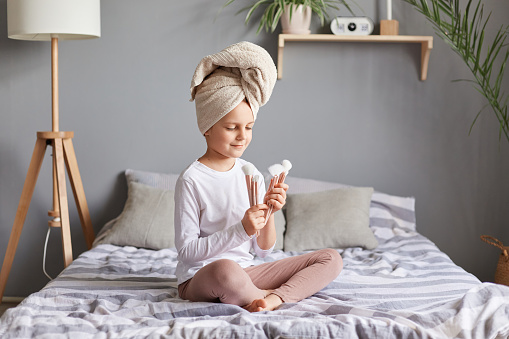 Cute little girl sitting on the bed holding set of makeup powder brushes, wearing white shirt and towel over head playing in bedroom after taking shower.