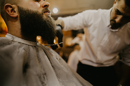 Close-up of a barber's hand with scissors, styling a client´s beard at the barber shop.