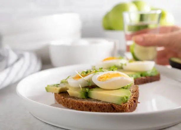 Delicious and healthy avocado egg sandwich with rye bread. Served on light kitchen counter  on a plate with glass of water. Closeup and front view