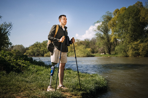 Hiking man with one artificial leg stands by a river in the forest