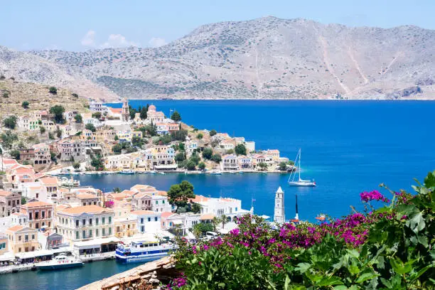 Photo of Picturesque view on tiny colorful houses on rocks and boats through blooming plants on balcony near the Mediterranian sea on Greek island in sunny summer day, vacation on exotic islands