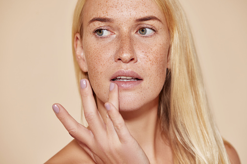 Woman with freckles touching her lip with a finger. Blond female with perfect skin applying balm on her lips.
