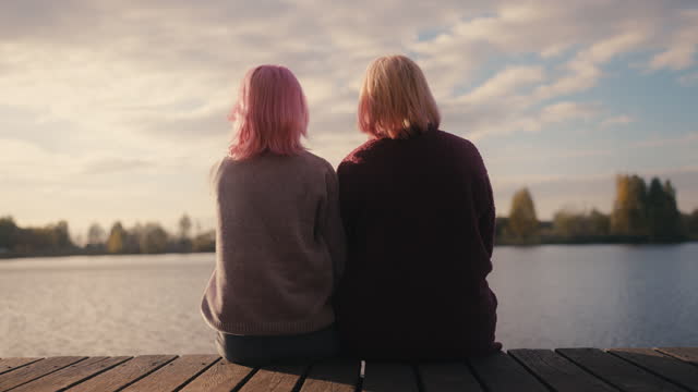 Back view of pink-haired daughter and mother sitting on wooden pierce by river