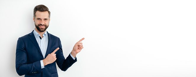 Panoramic photo of positive caucasian man, in elegant suit, pointing aside with fingers hands gesture at empty space for presentation or advertising, looks at camera, smiles. Isolated white background