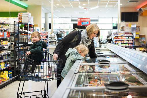 A woman leaning into a freezer with some help from her young daughter while out shopping for groceries in her local supermarket.