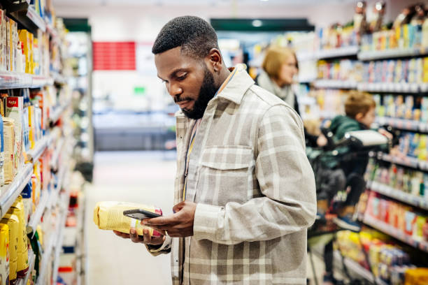 Man Looking Up Ingredients On Smartphone While Grocery Shopping A man using his smartphone and looking up ingredients from a product while grocery shopping in his local supermarket. smart phone technology lifestyles chain stock pictures, royalty-free photos & images