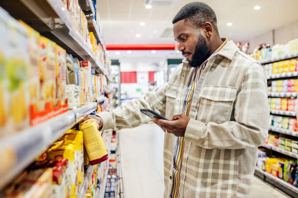 uomo che guarda lo smartphone mentre sceglie gli articoli nel supermercato - supermarket shopping retail choice foto e immagini stock