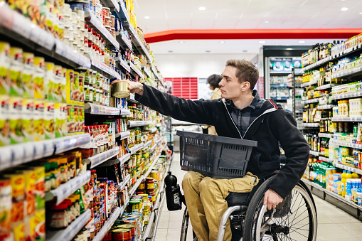 A man sitting in a wheelchair and with a basket on his lap, picking an item off a shelf while shopping for some groceries in his local supermarket.