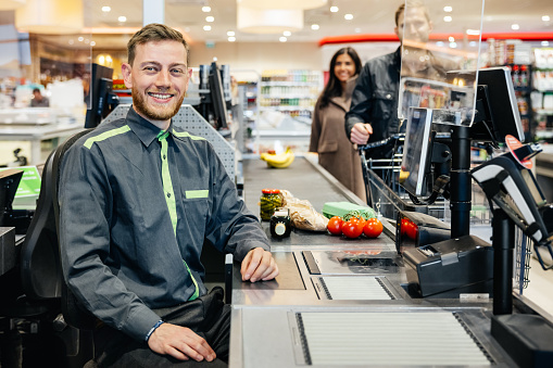 A portrait of a supermarket cashier sitting at his till, smiling while customers place their groceries son the conveyor.