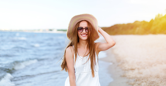 Happy smiling woman in free happiness bliss on ocean beach standing and posing with hat and sunglasses. Portrait of a female model in white summer dress enjoying nature.