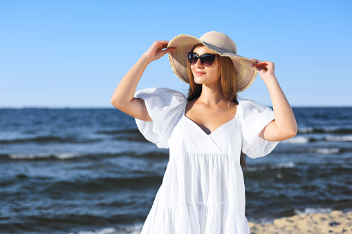 Happy blonde woman is posing on the ocean beach with sunglasses and a hat. Evening sun.