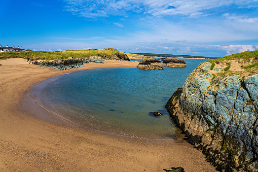 A beautiful small white beach in Scotland with azure blue water. There is a staircase between the grass and the rocks so that you can get to the beach. It is a cloudy day but the sun comes in occasionally.