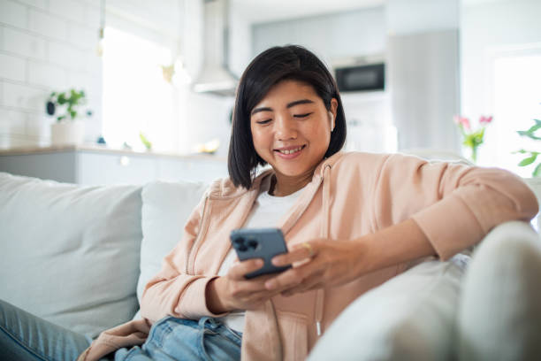 junge frau, die ein smartphone benutzt, während sie auf einer couch in einem wohnzimmer sitzt - op stock-fotos und bilder