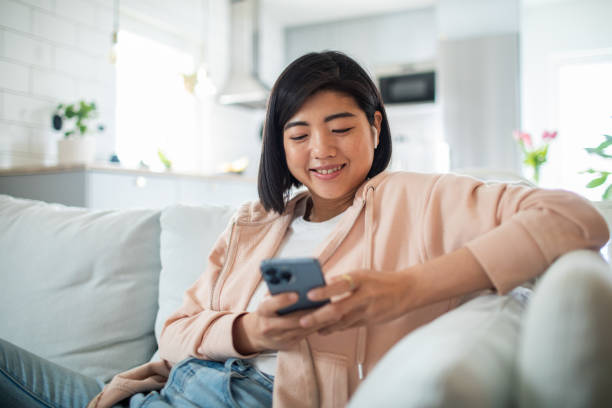mujer joven usando un teléfono inteligente mientras está sentada en un sofá en una sala de estar - usar el teléfono fotografías e imágenes de stock