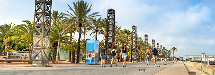 Barcelona, Spain - June 18, 2022:  People walk along the palm tree lined boardwalk by the hotels and outdoor restaurants on the Barceloneta Beach sand shore on the Balearic sea in Barcelona Catalonia Spain