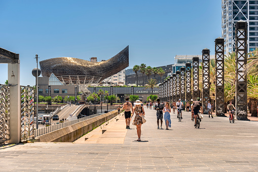 Barcelona, Spain - June 11, 2022:  Crowds of people relax and walk along the shore in the hot sun by the bars and restaurants on the Balearic sea and Barceloneta beach.  The city casino and Frank Gehry's modern Peix d'Or sculpture is on the boardwalk