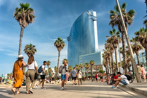 Barcelona, Spain - June 12, 2022:  People walk along the palm tree lined boardwalk by the hotels and outdoor restaurants on the Barceloneta Beach sand shore on the Balearic sea in Barcelona Catalonia Spain