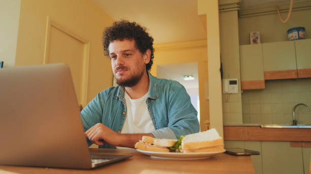istock Guy dressed in denim shirt works on laptop while having lunch in cozy kitchen 1485808234