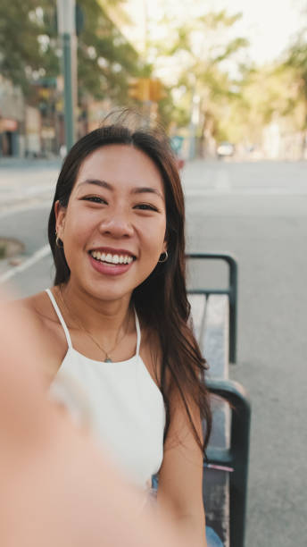 istock Pretty brunette girl dressed in white top and jeans is smiling making selfie while sitting on bench on cityscape background 1485809271