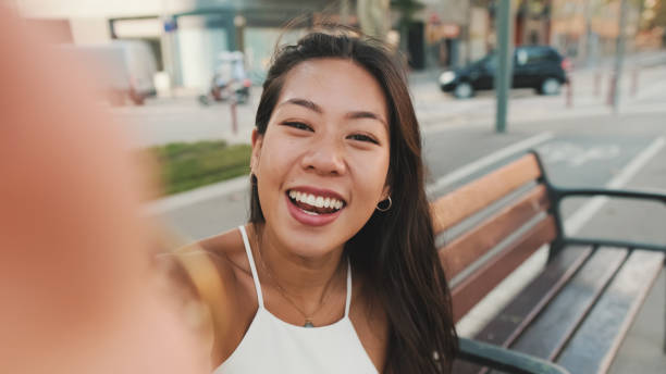 istock Pretty brunette girl dressed in white top and jeans is smiling making selfie while sitting on bench on cityscape background 1485809301