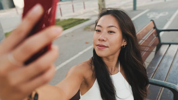 istock Pretty brunette girl dressed in white top and jeans is smiling making selfie while sitting on bench on cityscape background 1485809381