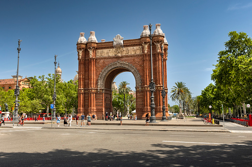 Barcelona, Spain - June 16, 2022:  People walk by the Arch of Triumph in the district of downtown Promenade Passeig de Lluís Companys.  In 1888 Barcelona hosted the Universal Exhibition. The Arc de Triomf was built as the gateway to the fair which was held in the Parc de la Ciutadella.