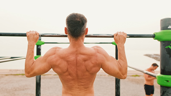 Middle-aged muscular man doing exercises on the horizontal bar on sea background, Back view