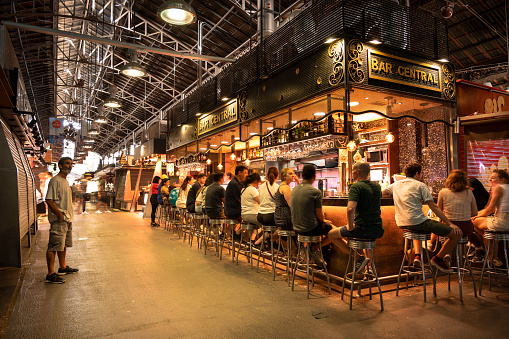 Barcelona, Spain - June 13, 2022:  People walk, shop and eat in the Mercado de La Boqueria open air food market by La Rambla where vendors sell meat, cheese and produce from the stalls and butcher counters