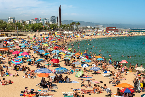 Barcelona, Spain - June 12, 2022:  Crowds of people relax and tan in the hot sun on the sand shores of the Balearic Sea on Barceloneta beach in Barcelona Catalonia Spain
