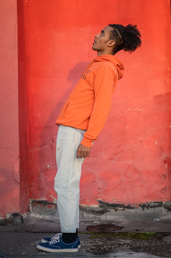 A side view of a man leaning backwards with his arms straight down at his sides, wearing casual trousers and a bright orange background
