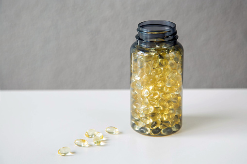 The transparent grey jar is on the table with scattered yellow transparent capsules with vitamin D or Omega-3 on a gray background. Copy space.