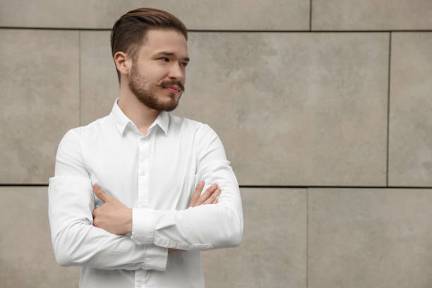 istock Smiling man in white shirt standing near light grey brick wall. Space for text 1485824633