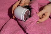 Woman using Anti-pilling razor at home. Device for shaving clothes. Anti-Plush fabric Shaver. Electric portable sweater pill defuzzer Lint remover from pink acrylic or wool sweater. Slow fashion mending repairing