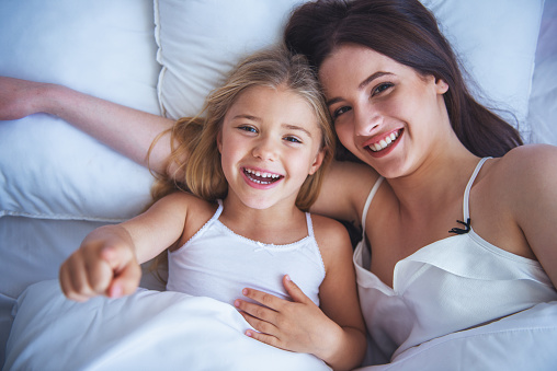 Top view of beautiful young mother and her daughter hugging, looking at camera and smiling while lying in bed at home