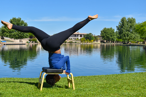 A beautiful, young African-American woman does yoga and stretches with her black yoga mat and headstand board in the grass on the shore of a manmade lake in southern Arizona on a sunny day wearing a blue activewear shirt with black leggings.