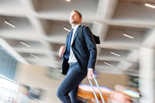 Caucasian business man wearing a dark suit and tie is walking at the baggage claim area of the airport and the background is in blurred motion.