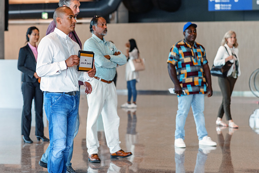 Multiracial people standing and waiting for their relatives and clients to arrive at the arrival zone of the airport. There is one Latin driver holding an Ipad with the name of his clients to take them to their hotel.