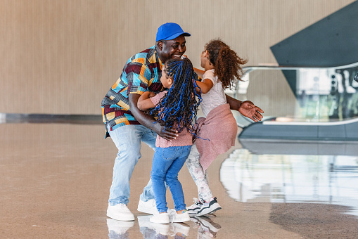 Two dark-skinned girls are happy to see their father at the arrival zone at the airport. His father is giving a hug to them and smiling.