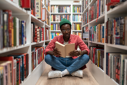 Student african american man reading research textbook in university library sitting on floor between bookshelf. Thoughtful guy hipster studying in public place. Education, learning, researching.