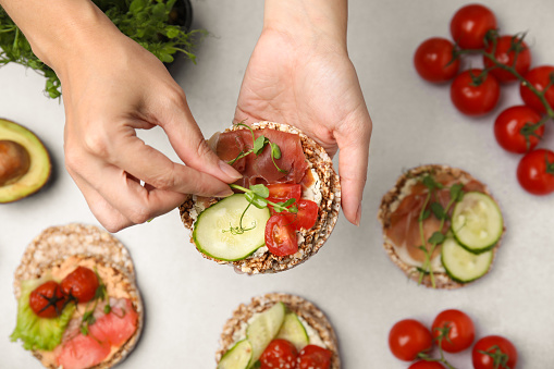 Woman adding microgreens onto crunchy buckwheat cakes with prosciutto, pieces of tomato and cucumber slice at white table, closeup