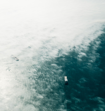 Taken from the window of a Cessna 172 over the San Pedro Harbor, California. Clouds and fog from the marine layer coat the sky. Huge cargo/container ships in the ocean play peekaboo through the white puffy fog. Two cargo ships are visible, one exposed and the other under a layer of haze. The ocean is a beautiful peacock shade of blue. Ripples from the wind and tide can be seen, outlined by the sunlight.