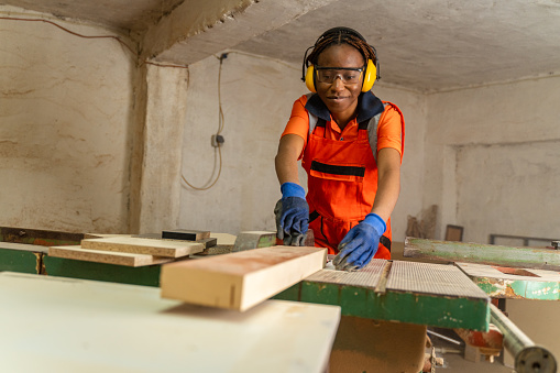 Female worker wearing ear and eye protectors while working. Carpenter is cutting wood with circular saw on workbench. She is making furniture in workshop.