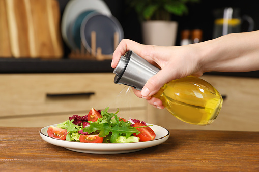 Woman spraying cooking oil onto salad on wooden table in kitchen, closeup
