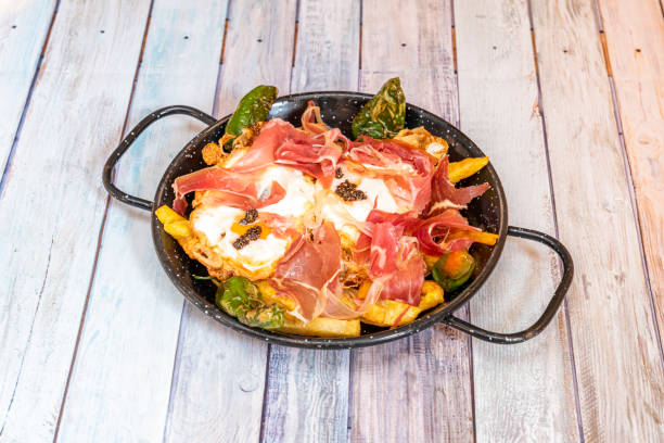 Delicious typical Spanish dish of broken eggs with serrano ham, fried peppers, homemade chips and truffle in a black enameled pan