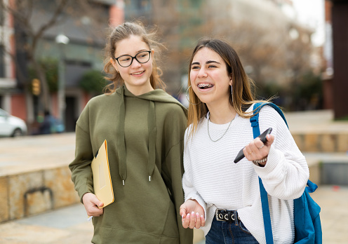 Two smiling teenager girls friends walking and laughing outdoors in city on a sunny day