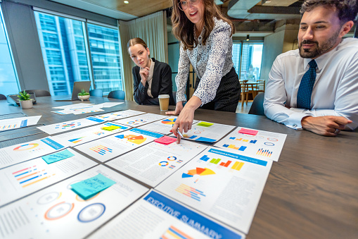 Paperwork and hands on a board room table at a business presentation or seminar. The documents have financial or marketing figures, graphs and charts on them. A woman is pointing at the data